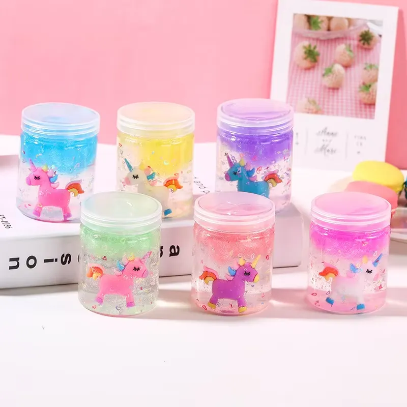 Kit de Slime Transparente para Niños – seis frascos de slime pastel con amuletos de unicornio dispuestos sobre un libro junto a una bolsa de bolas de espuma multicolores.