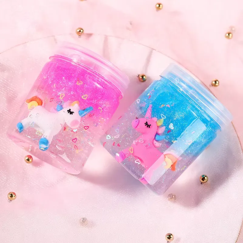 Kit de Slime Transparente para Niños – dos frascos de slime pastel tumbados de lado sobre tela rosa, revelando amuletos de unicornio y confeti de corazones flotantes.